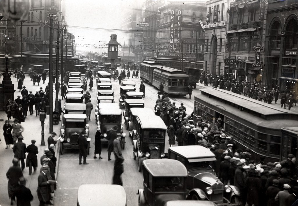 Corner of Michigan and Griswold. Great deal of car traffic, large group of people boarding trolley car. Large commercial buildings in background. Traffic tower in middle of street, with person standing inside. Date 	  circa 1920 Source 	  Early Detroit Images from the Burton Historical Collection, Detroit Public Library Author 	  unknown
